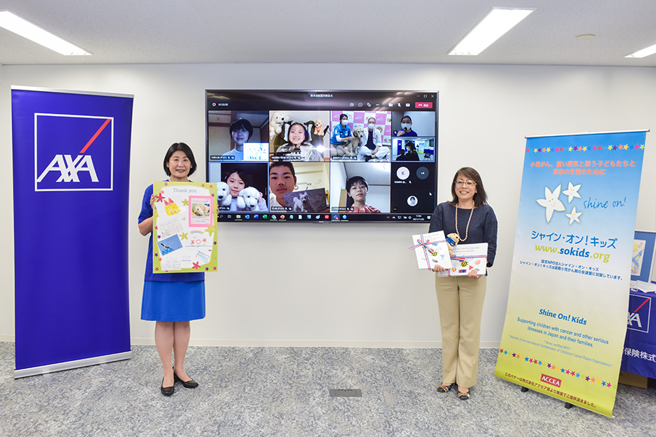 「スマイルランドのミツバチ〜新しい世界へ〜」完成お披露目会および贈呈式 シャイン・オン!キッズ 事務局長 ニーリー美穂氏(右) アクサ損害保険株式会社 代表取締役社長兼CEO 佐伯美奈子(左) モニター画面には神奈川県立こども医療センター病院 総長 町田治郎氏(画面上右から2人目)と、シャイン・オン!キッズ ハンドラー 森田優子氏(画面上右から3人目)、アニー、絵本作者の子どもたち